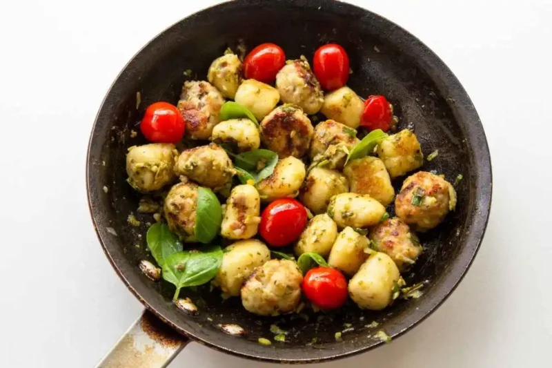 Pan-seared Tuscan chicken meatballs paired with gnocchi, cherry tomatoes, and fresh basil, garnished with pesto in a skillet.