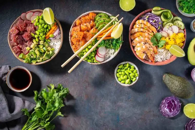 A variety of colorful poke bowls with tuna, salmon, shrimp, vegetables, and rice, garnished with lime, edamame, and fresh herbs.