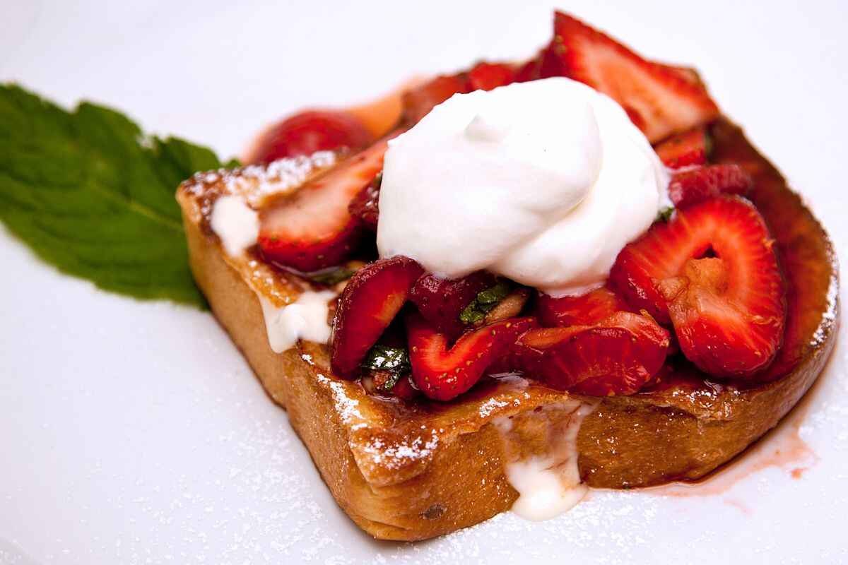 A close-up of brioche French toast casserole topped with fresh strawberries, a dollop of whipped cream, and dusted with powdered sugar, served on a white plate with a mint leaf garnish.