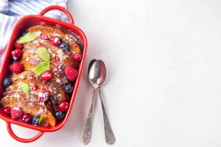 A vibrant brioche French toast casserole in a red baking dish, topped with fresh berries, mint leaves, and a sprinkle of powdered sugar, styled with silver spoons on a light background.
