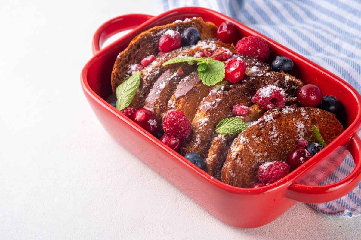 Side view of a brioche French toast casserole layered with slices of golden-baked brioche, fresh berries, and garnished with mint leaves, served in a bright red dish with a striped kitchen towel.