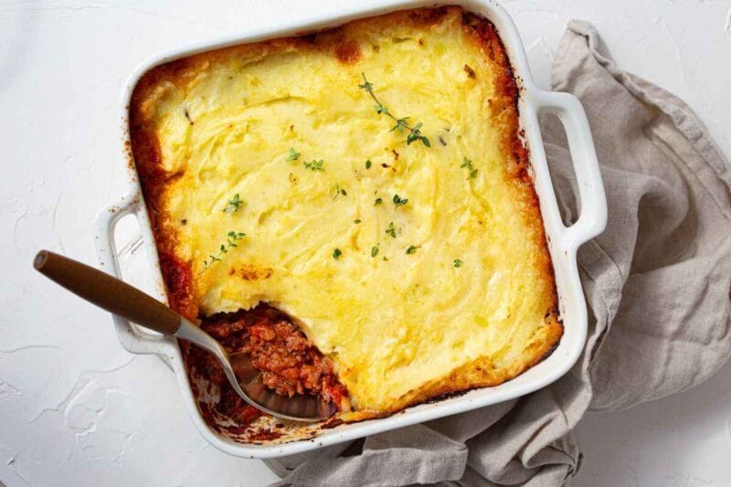 A rich Casserole Dish with creamy mashed potato topping in a white dish, garnished with fresh thyme, with a serving spoon revealing the savory meat filling underneath.