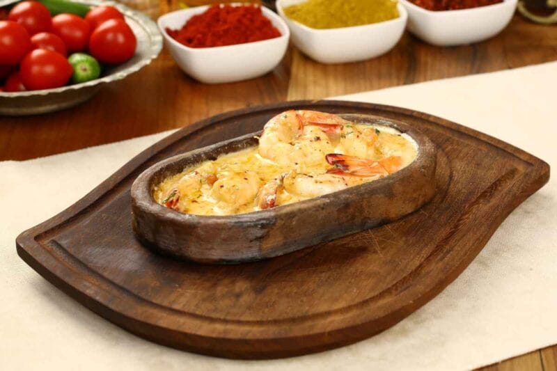 A rustic shrimp casserole served in a sizzling hot dish, featuring juicy shrimp in a creamy, spiced sauce, placed on a wooden platter with fresh tomatoes and spices in small bowls in the background.