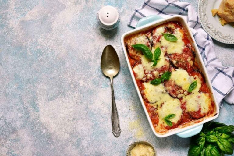 Top-down view of a cheesy eggplant casserole baked to perfection, garnished with fresh basil leaves, styled on a rustic blue surface with a spoon and small bowl of grated parmesan cheese nearby.