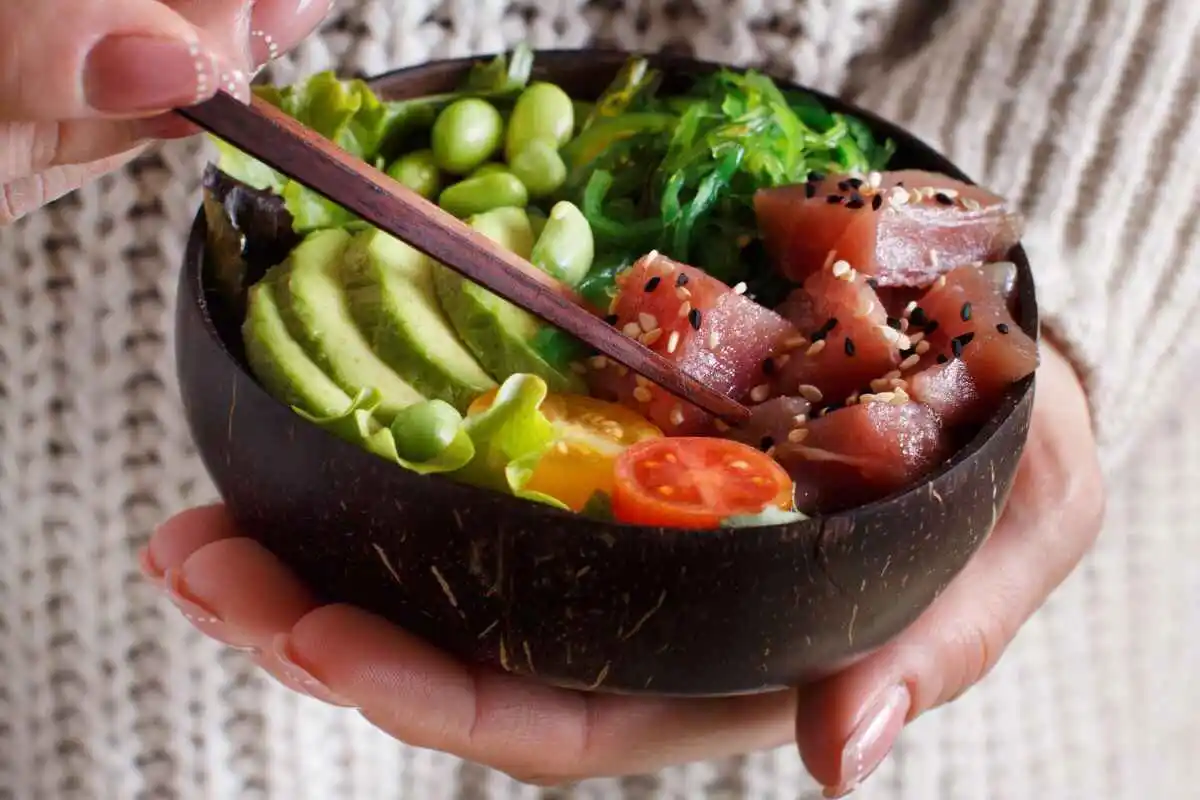A colorful poke bowl filled with tuna, avocado, edamame, cherry tomatoes, and seaweed salad, topped with sesame seeds in a coconut bowl.