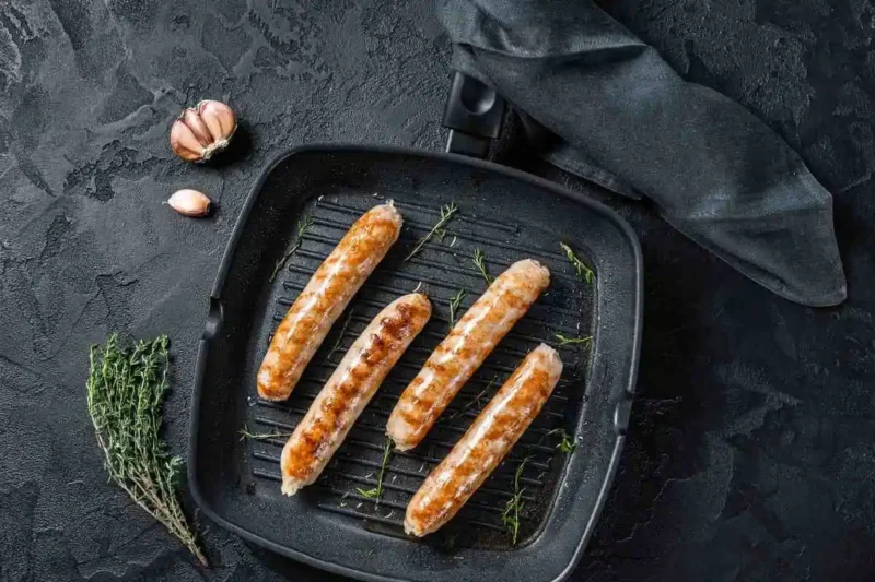 Four grilled chicken sausages in a grill pan, accompanied by sprigs of fresh thyme and garlic, ready for a healthy and delicious meal.