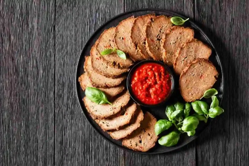 Sliced meatloaf served on a black plate with fresh basil and a side of marinara sauce, perfect for a hearty and flavorful meal."