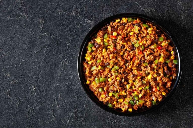 A hearty Mexican Corn casserole with ground meat, black beans, corn, and green peppers, presented in a black dish against a textured dark gray background for a bold and appetizing look.