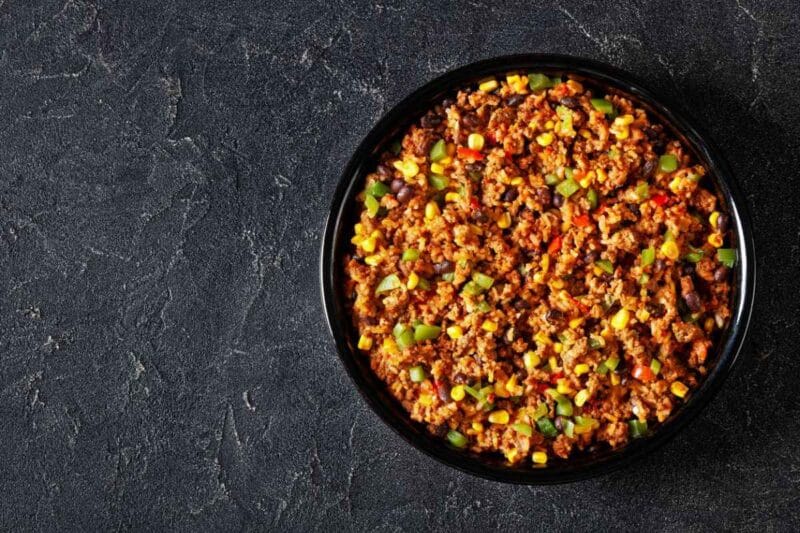 A hearty Mexican Corn casserole with ground meat, black beans, corn, and green peppers, presented in a black dish against a textured dark gray background for a bold and appetizing look.