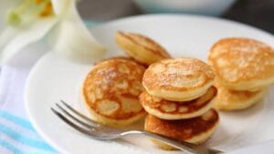 A plate of golden-brown mini pancakes dusted with powdered sugar, served with a fork on a white plate.