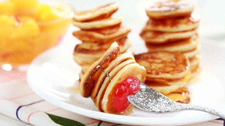 Stacks of golden mini pancakes served on a white plate, topped with powdered sugar and a cherry on a fork.