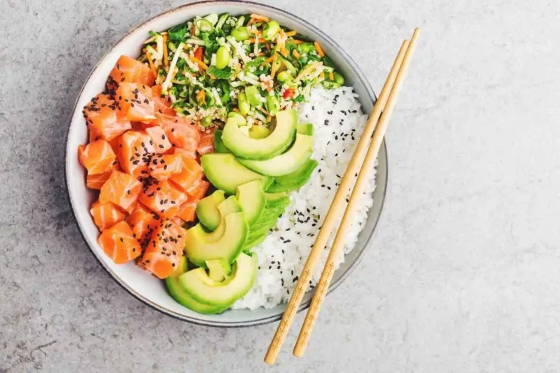 A fresh poke bowl with diced salmon, avocado slices, quinoa salad, and rice, garnished with black sesame seeds, served in a white bowl