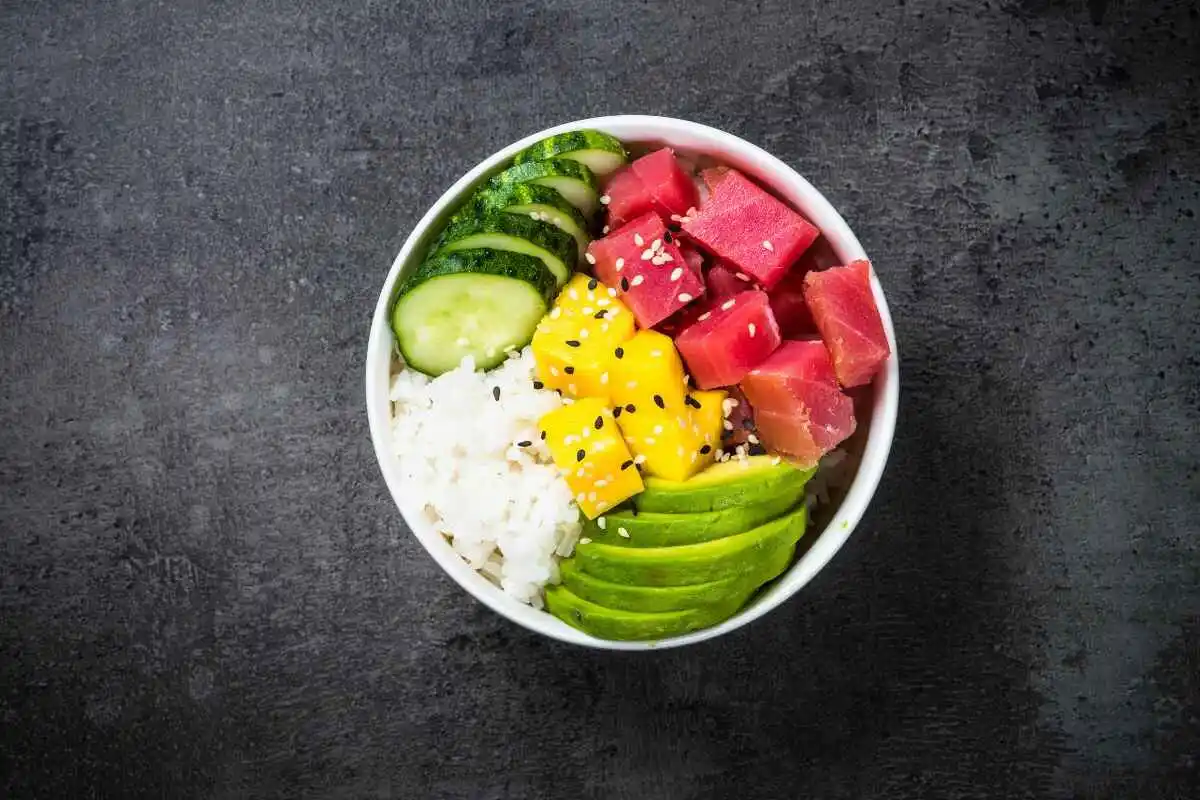 Is Poke Eaten Cold or Hot? A colorful poke bowl with diced tuna, mango, cucumber, avocado slices, and rice, topped with sesame seeds, served in a white bowl.