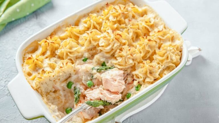 A freshly baked salmon casserole with golden, piped mashed potato topping, mixed with asparagus, peas, and creamy sauce, served in a green and white baking dish.