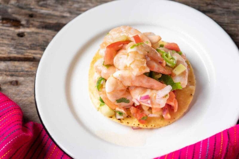 Close-up view of a vibrant Shrimp Tostadas served on a white plate, topped with juicy shrimp, fresh tomatoes, onions, cilantro, and jalapeño slices, set on a rustic wooden table with a colorful pink napkin.
