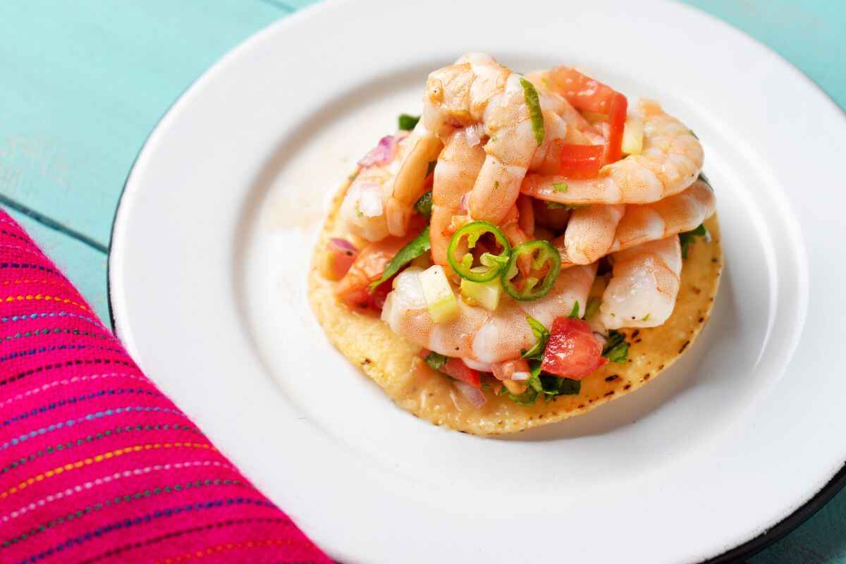 A beautifully plated Shrimp Tostadas featuring plump shrimp, fresh vegetables, and a crispy tortilla, styled against a light turquoise background with a bright pink napkin adding a pop of color.
