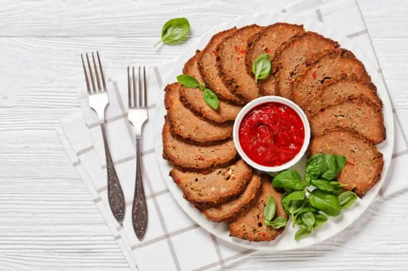Slices of smoked meatloaf arranged on a white plate, served with marinara sauce, fresh basil, and two forks for a classic dinner setup.
