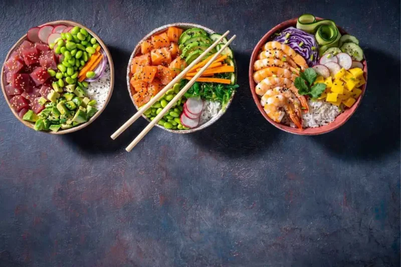 Three vibrant poke bowls featuring tuna, salmon, shrimp, fresh vegetables, and rice, beautifully garnished and served with chopsticks.