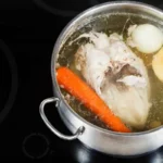 A pot of boiling chicken with vegetables, including carrots, onions, and a whole chicken, simmering in a broth on a stovetop.