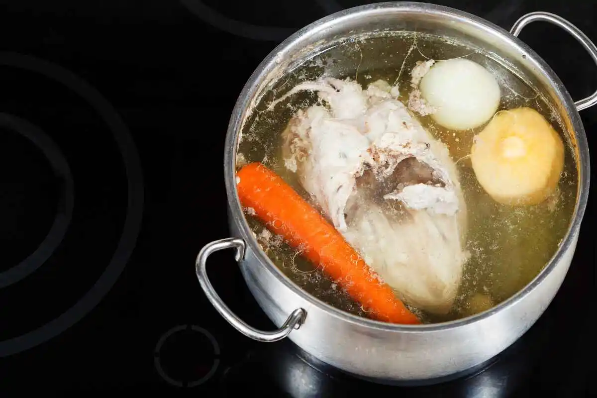 A pot of boiling chicken with vegetables, including carrots, onions, and a whole chicken, simmering in a broth on a stovetop.