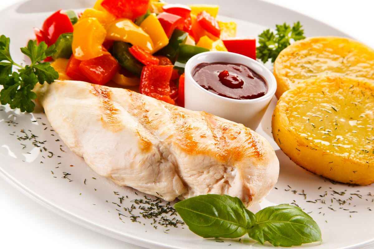 Is a Turkey a Type of Chicken? Grilled chicken breast served with a colorful vegetable salad, a side of round, seasoned potatoes, and a small dish of dipping sauce on a white plate.