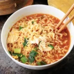 A bowl of homemade ramen noodles with a spicy broth, egg pieces, and chopped green onions, with a pair of chopsticks picking up some noodles.