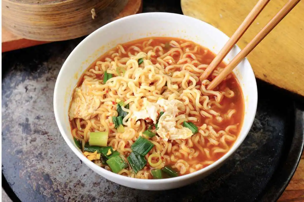 A bowl of homemade ramen noodles with a spicy broth, egg pieces, and chopped green onions, with a pair of chopsticks picking up some noodles.