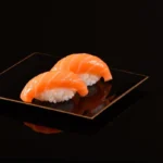 Two pieces of salmon nigiri sushi, artistically displayed on a sleek black plate with a subtle golden edge, reflecting a minimalist and modern aesthetic.