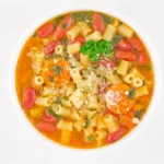 A colorful bowl of pasta e fagioli soup, featuring ditalini pasta, kidney beans, carrots, and a rich tomato broth garnished with parsley and Parmesan cheese, captured from an overhead view.