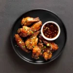 Smoked chicken wings garnished with green onions, served with a bowl of dipping sauce on a black plate.