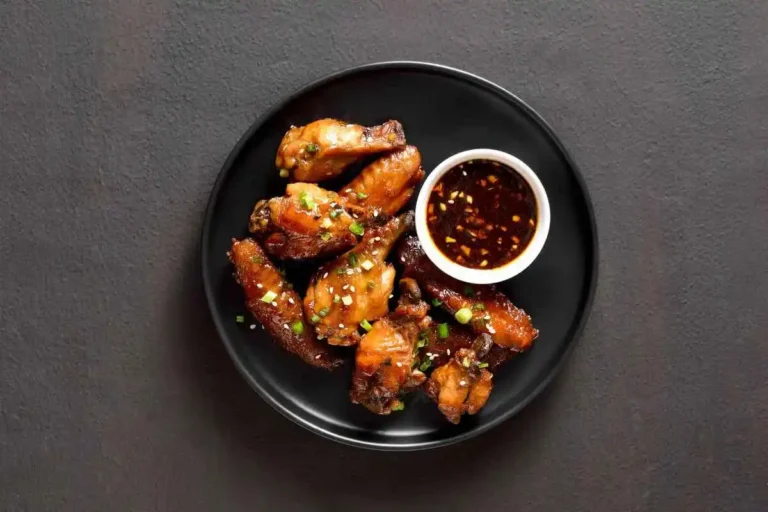 Smoked chicken wings garnished with green onions, served with a bowl of dipping sauce on a black plate.