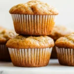 A stack of freshly baked banana bread muffins with a golden-brown crust, set on a white surface with additional muffins in the background.