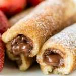 Close-up of Nutella-filled French toast roll-ups coated in cinnamon sugar, with melted chocolate oozing out, served with fresh strawberries.