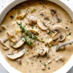 A bowl of rich and creamy mushroom soup garnished with fresh thyme and black pepper, served with a spoon.